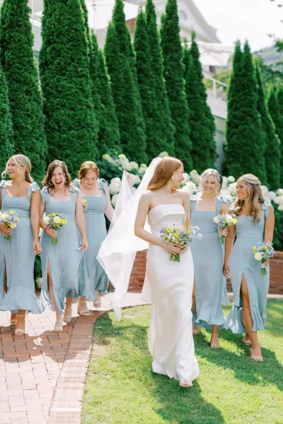 Bride with bridesmaids walking together in dusty blue dresses, the bride in a strapless gown with veil blowing on a garden lawn