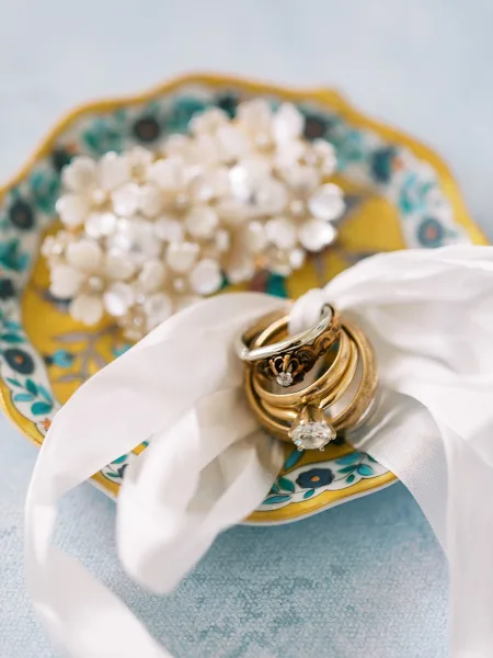 Wedding rings arranged in a wedding ring flat lay with diamond ring and gold bands tied with white ribbon on a floral plate on light blue surface