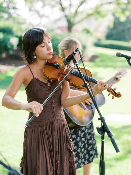 Wedding musicians perform live with a violin and acoustic guitar at microphones on a green garden lawn with trees in the background