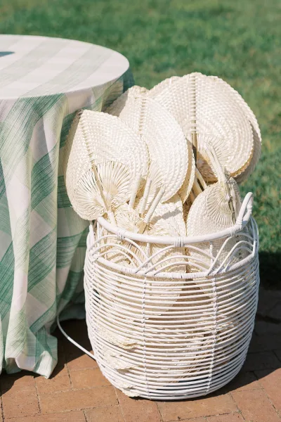 Wedding fan basket filled with woven hand fans in a white wicker basket on a green plaid tablecloth beside a brick patio lawn