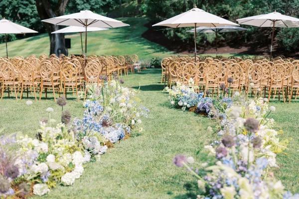 Ceremony aisle decor with hydrangea and delphinium florals lining a curved outdoor ceremony aisle, wood chairs and white umbrellas on lawn