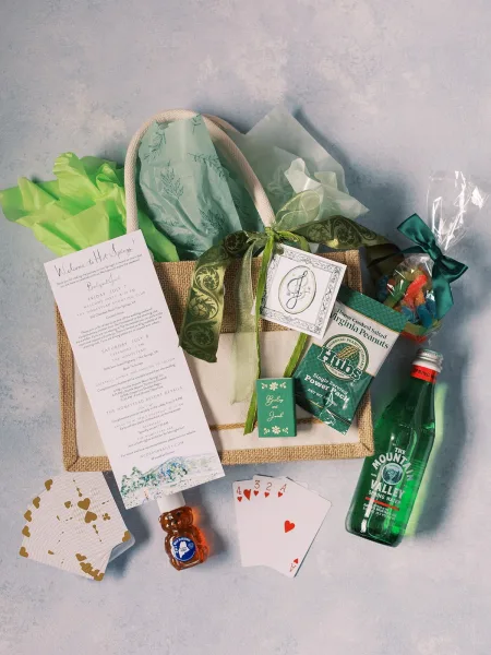 Welcome bag flat lay with jute tote, tissue paper and ribbon bow, gift tag, itinerary card, snacks, water, candy, playing cards on gray backdrop