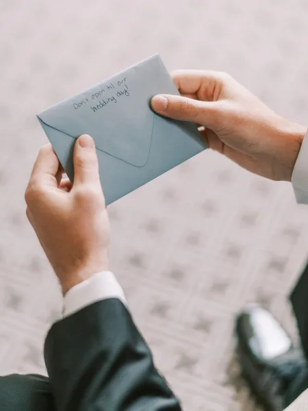Wedding letter held by a groom reading letter, showing a blue envelope and handwritten note beside suit cuffs on patterned carpet floor