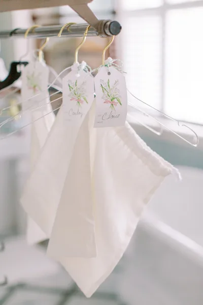 Wedding hanger tags and bridesmaid dress hanger tags with floral illustration and ribbon tied to hangers on a clothing rack by window blinds