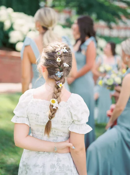 Flower girl hairstyle with a flower girl braid and tiny blooms woven in, standing in a garden bridal party with bouquets by a brick wall