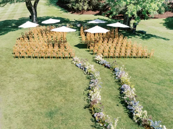 Ceremony setup with wood chairs and white umbrellas framing a curved aisle lined with pastel florals on a grassy garden lawn