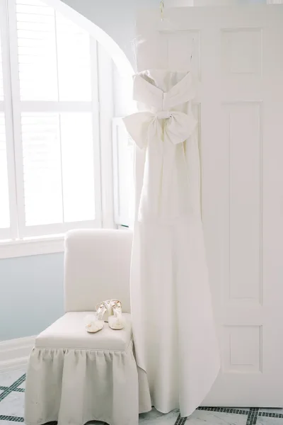 Wedding dress, strapless wedding dress with oversized bow hanging on a white door above bridal shoes on an upholstered chair