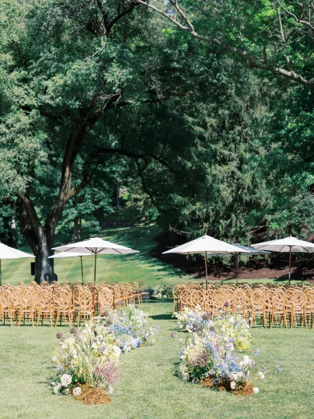 Ceremony setup with wood chairs lining a lawn aisle, blue and white floral clusters, and white umbrellas under trees on a hillside lawn