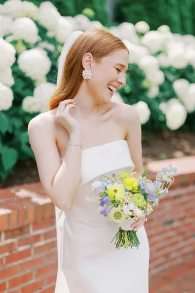 Bridal portrait of a laughing bride in a strapless wedding dress holding a colorful wildflower bouquet before hydrangeas and brick wall