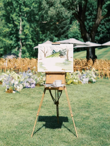 Wedding live painter creating a live wedding painting on an easel beside ceremony chairs and white umbrellas on a grassy lawn, palette in hand