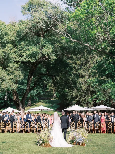 Outdoor wedding ceremony with a bride walking down the aisle, long veil and train, toward a floral arch on a green garden lawn