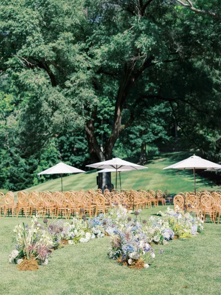 Ceremony setup with outdoor ceremony chairs lining a grass aisle, low blue-and-white florals, and white patio umbrellas beneath trees