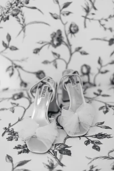 Bridal shoes in a wedding shoes flat lay, white heeled sandals with tulle bow accents and ankle straps on a floral bedspread
