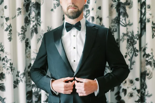 Groom portrait in a black tuxedo look with bow tie, white shirt studs and cufflinks, posed indoors by patterned curtains