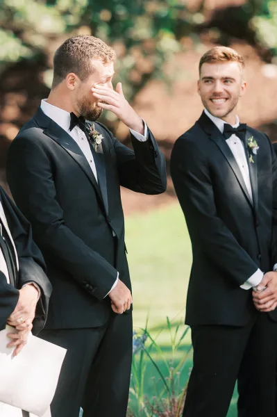 Groom crying as he wipes away tears, in a black tuxedo with boutonniere, beside smiling groomsmen against outdoor greenery