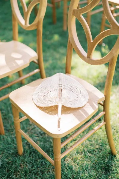 Wedding ceremony seating with outdoor ceremony chairs on a grass lawn, featuring wood chiavari chair backs and a woven hand fan on each seat