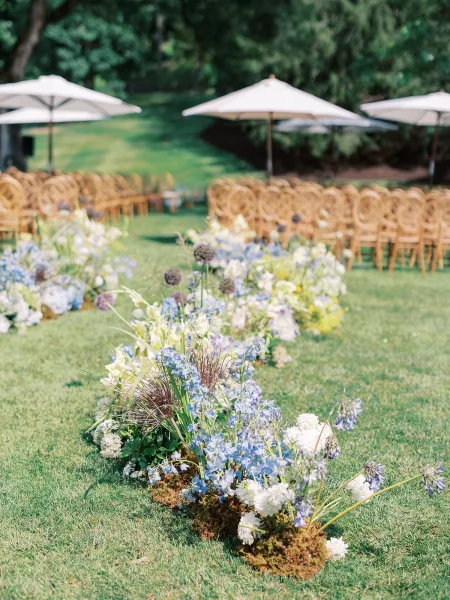 Ceremony aisle decor with meadow aisle flowers in blue and white, moss and ground florals bordering wicker chairs on a garden lawn