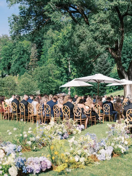 Outdoor ceremony seating with wood chairs and white patio umbrellas lining a floral aisle on a lawn beneath tall trees and blue sky