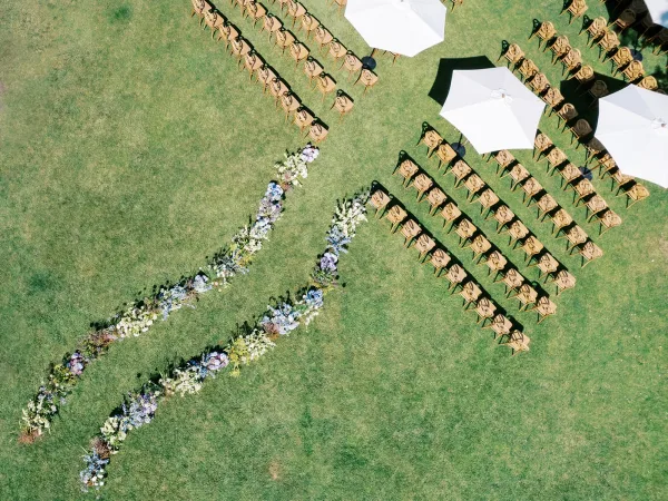 Ceremony aisle design with floral aisle runner and wood ceremony chairs, lined with pastel ground flowers beneath white umbrellas on a green lawn