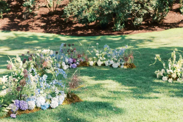 Ceremony aisle florals with meadow aisle flowers—low hydrangea, delphinium and allium clusters on moss along a sunlit grass lawn aisle