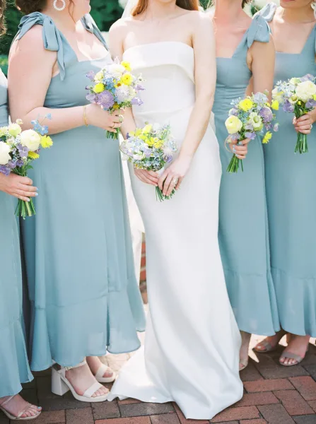 Bridesmaid group photo with dusty blue bridesmaid dresses and the bride in strapless white, holding pastel bouquets on a brick patio backdrop