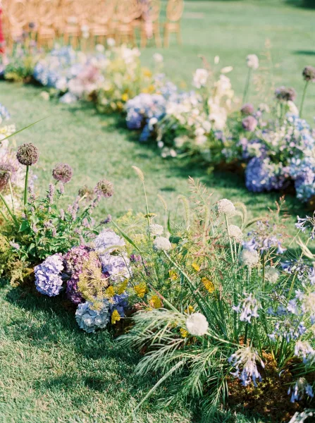 Ceremony aisle florals with grounded aisle flowers of blue hydrangeas and wildflowers lining a grass lawn aisle beside chairs