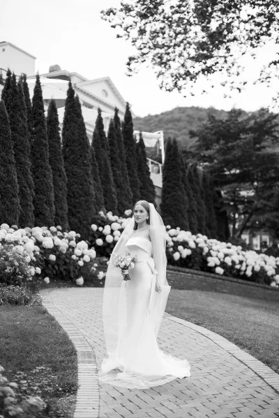 Bridal portrait of a bride holding bouquet in a strapless gown with long veil and drop earrings on a garden path by hydrangeas and mountains