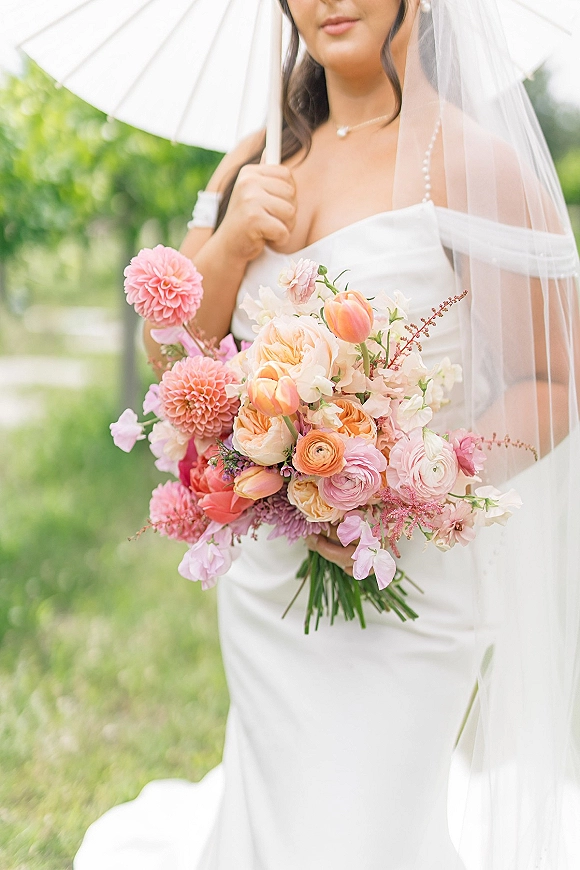 Bridal bouquet, pink and peach bouquet held by a bride in a strapless dress with veil and pearl necklace, under a white parasol outdoors
