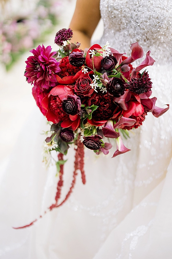 Bridal bouquet of deep red blooms with calla lilies, roses, and cascading greenery against a beaded wedding dress and soft white backdrop