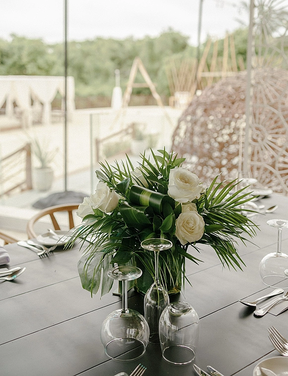 Reception centerpiece with white rose centerpiece and palm fronds in a glass vase on a black table with wine glasses on an outdoor patio