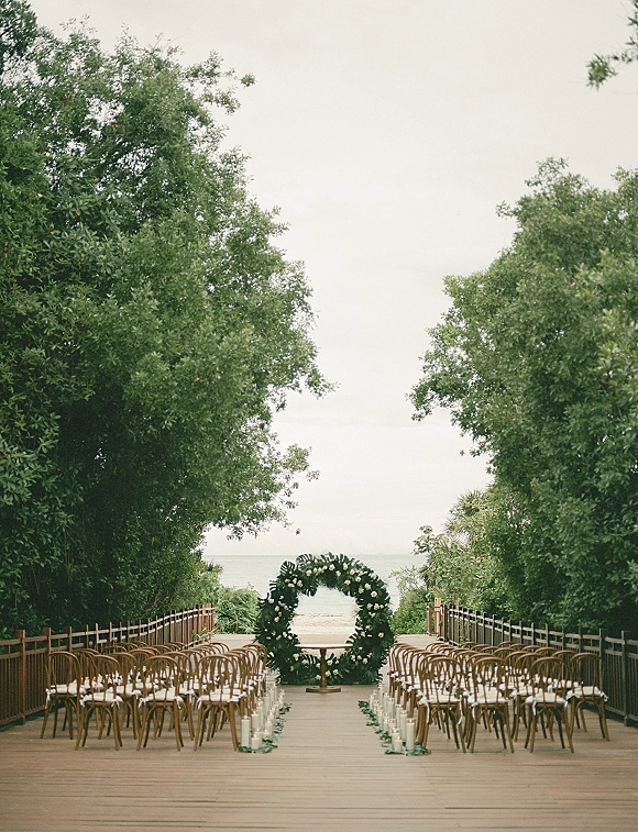 Ceremony setup with an outdoor wedding aisle lined with glass candle holders, wooden chairs, and a circular floral arch facing the ocean