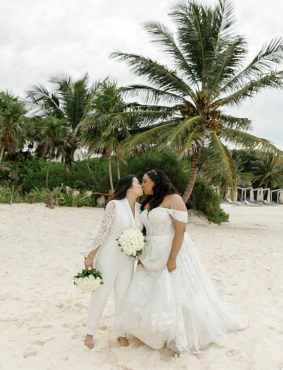 Wedding kiss portrait of two brides kissing on a sandy beach, holding white rose bouquets with palm trees and tropical greenery behind them