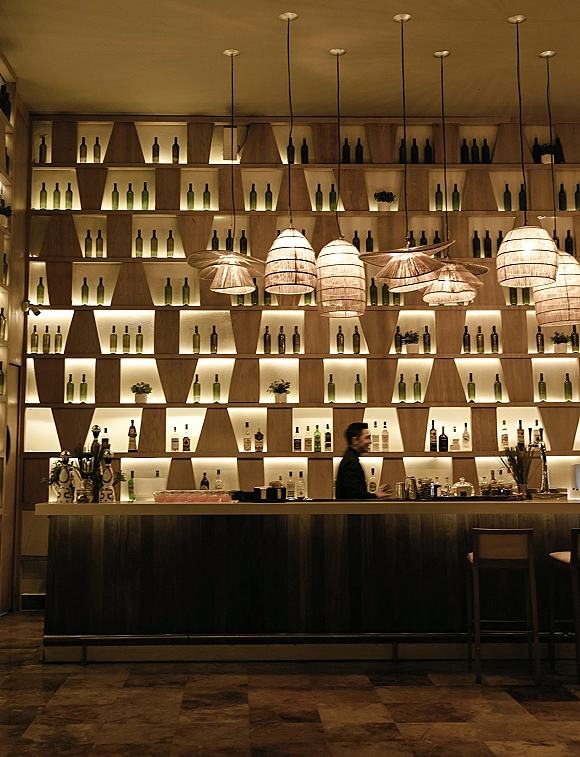 Wedding bar setup with backlit bottle display on wooden shelving, pendant lanterns over the counter, glassware, stools, and floral accent indoors