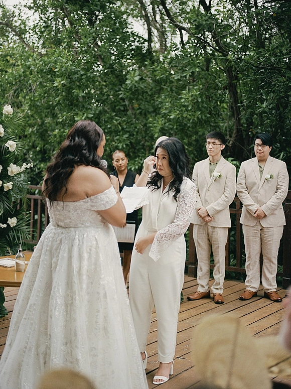 Wedding vows as bride reading vows into a microphone, off-the-shoulder lace sleeves visible on an outdoor deck amid lush greenery