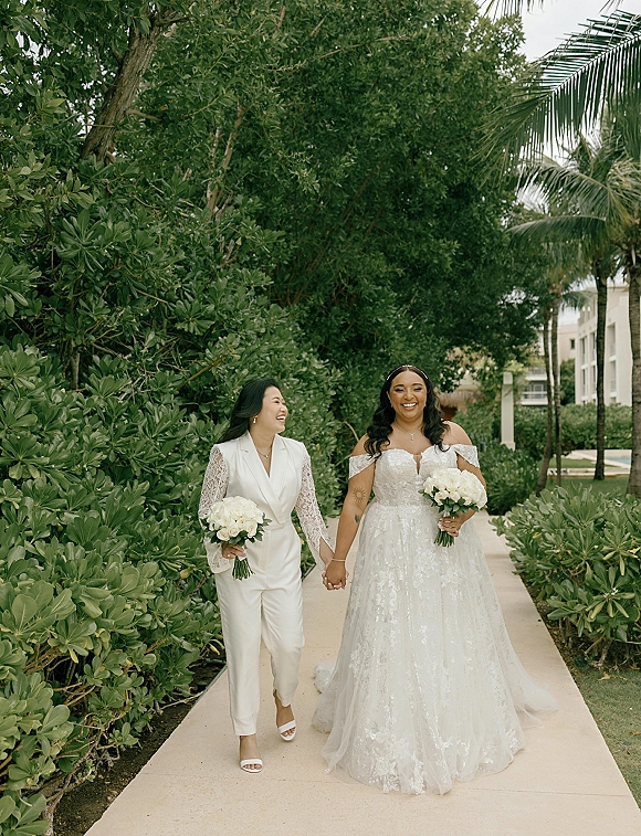 Couple portrait of two brides holding hands and walking, one in a white suit, the other in an off-shoulder lace gown with veil, in a lush garden walkway