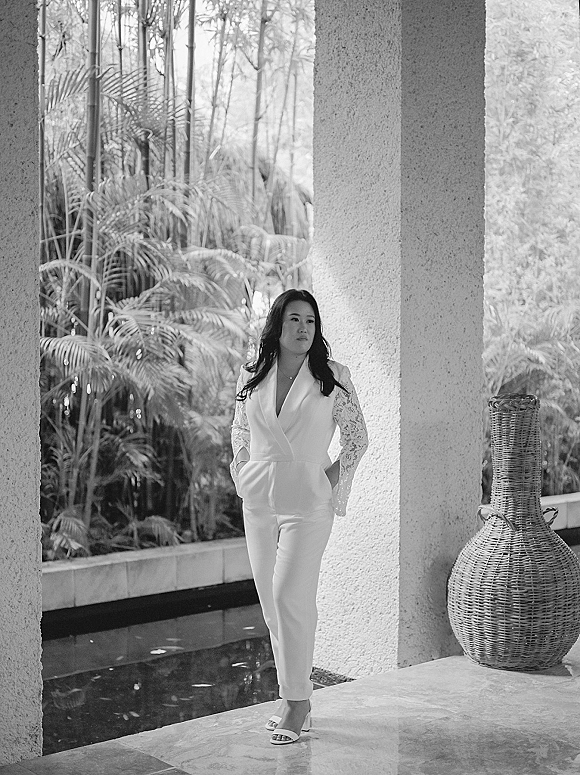 Bridal portrait of a bride in a bridal jumpsuit with lace sleeves and necklace, standing by concrete columns near a reflecting pool