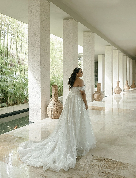 Bridal portrait of a bride in an off the shoulder wedding dress with chapel train, looking back in a stone colonnade by a reflecting pool