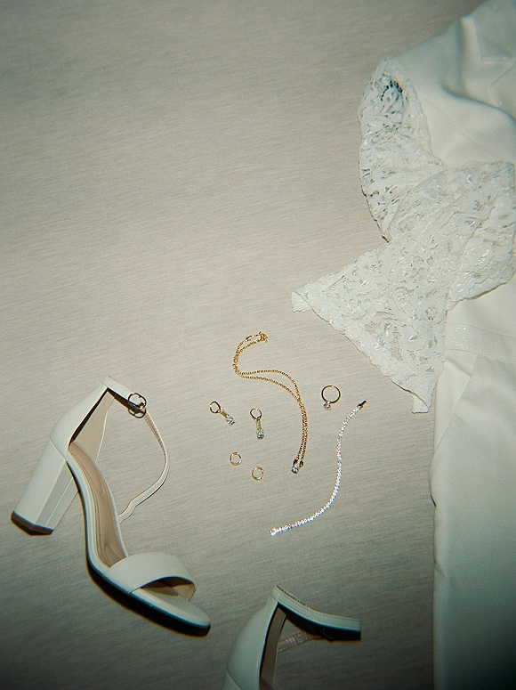 Bridal accessories flatlay with wedding jewelry flat lay, white heeled sandals, lace bodice, rings, earrings, and gold necklace on neutral fabric surface