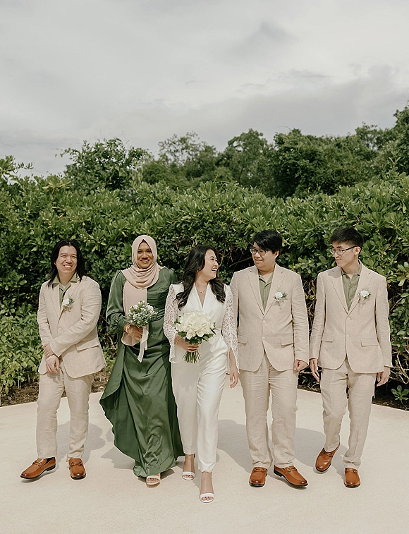 Wedding party portrait with bride in a white jumpsuit and bridesmaids and groomsmen walking, laughing by green hedges under a cloudy sky