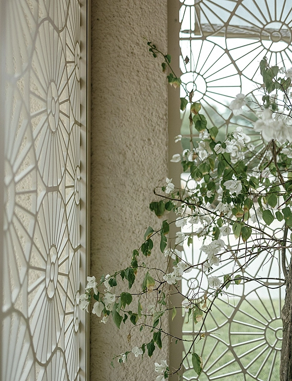 Ceremony backdrop with white flowers and greenery vines framing a decorative window screen on a stucco wall, accented by branches