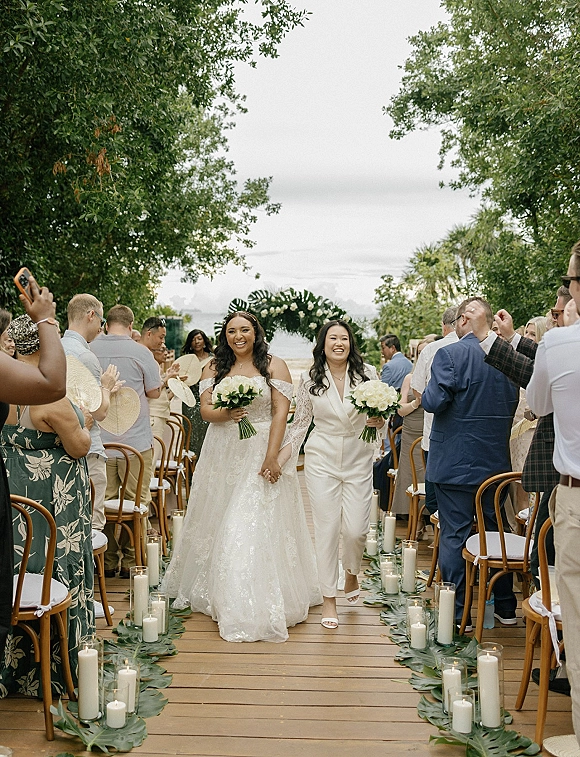 Wedding recessional as two brides hold hands and walk the candle-lined outdoor aisle, carrying white bouquets beneath a floral arch