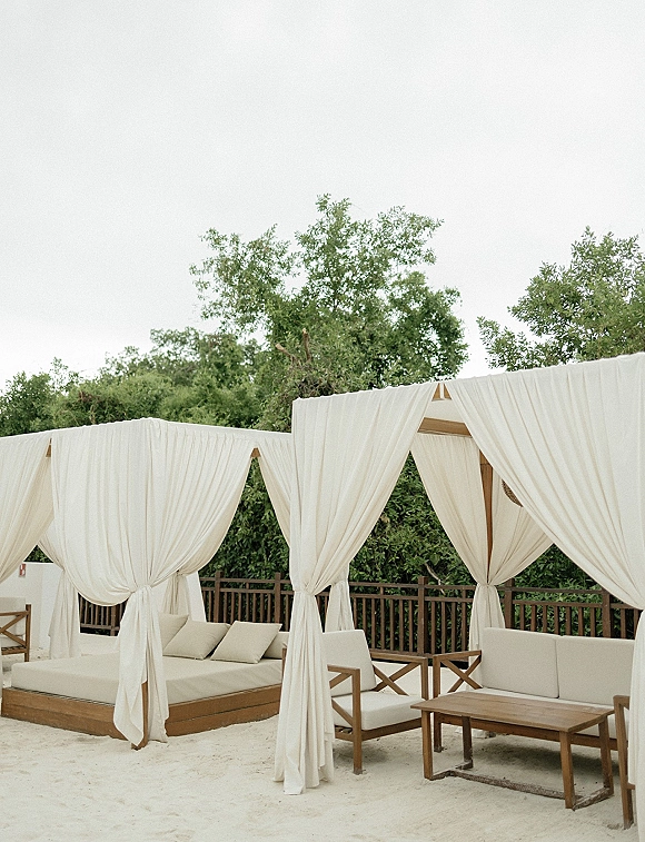 Wedding lounge setup with outdoor wedding lounge cabana daybeds, white drapery, sofas and pillows around a wooden coffee table on sand