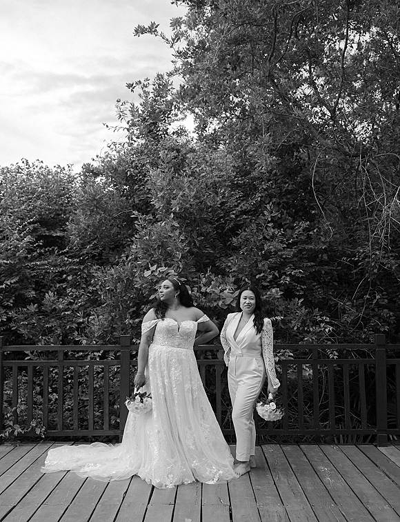 Bride and bride portrait of two brides holding matching white rose bouquets, one in a lace-sleeved jumpsuit, on a wooden deck with trees behind