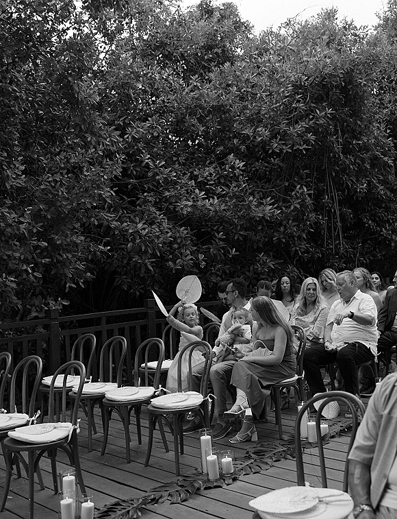 Ceremony guests seated in wedding guest seating on an outdoor deck, facing a candle-lined greenery aisle with wooden railing and trees beyond