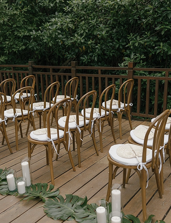 Ceremony seating with bentwood wedding chairs, white cushions and ribbon ties, woven fans, and a candle-lined aisle on a wood deck