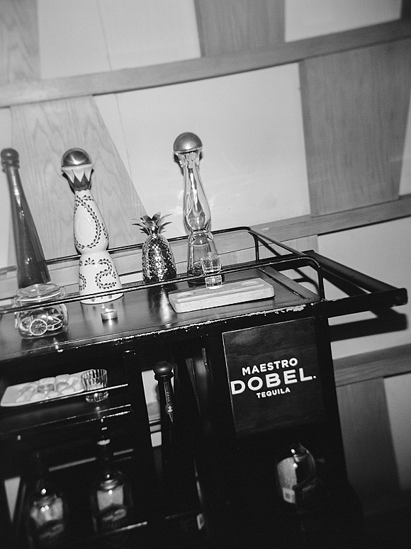 Tequila bar setup on a bar cart with tequila bottle, shot glasses, pineapple decor, dried citrus jar, and candle against wood wall shelving