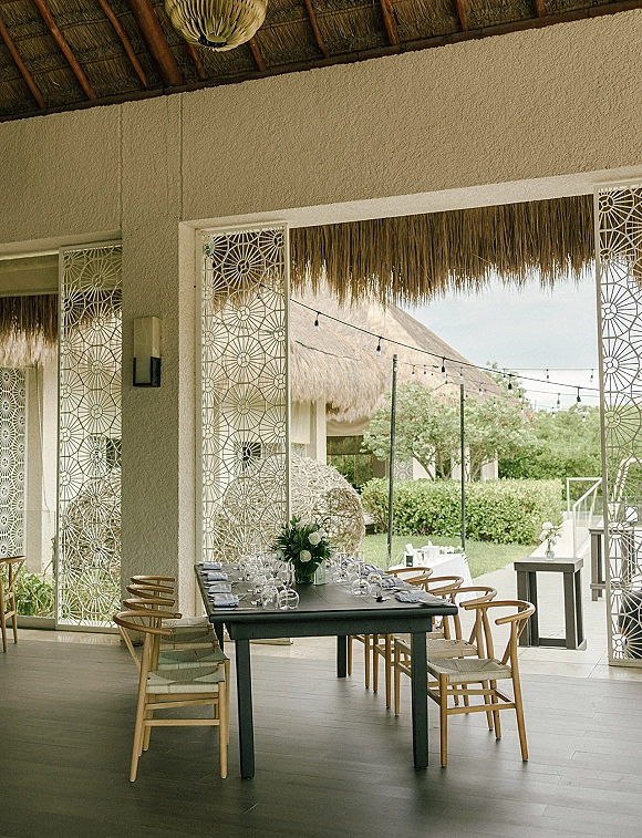 Reception tablescape with an outdoor reception table set with white floral centerpiece, glassware, and string lights on a covered patio under a thatched roof