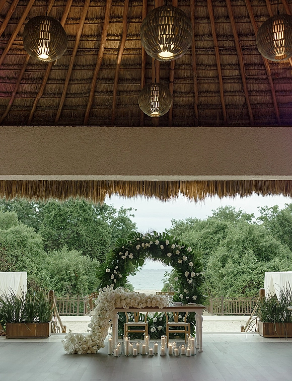 Wedding ceremony setup with circular floral arch of white roses and tropical greenery, candlelit altar under a thatched roof by the ocean view