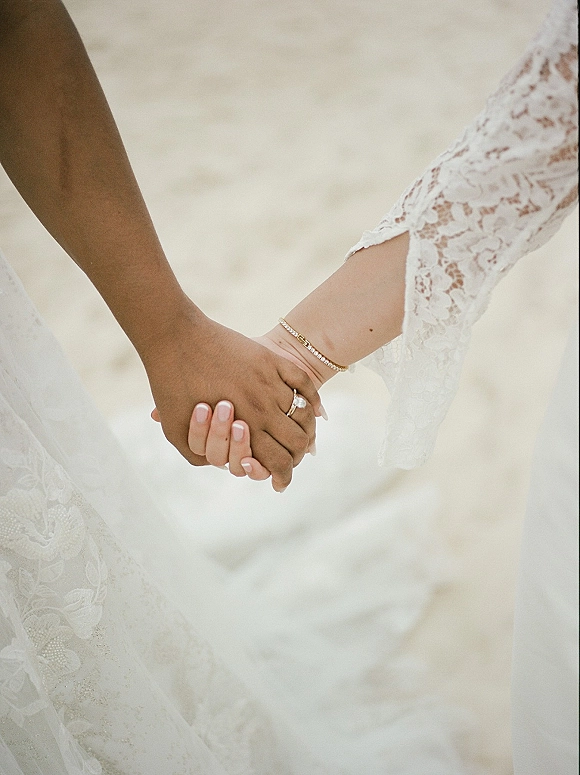 Wedding hand holding with wedding rings in close up, showing lace sleeve, bracelet and french manicure against beach sand shoreline