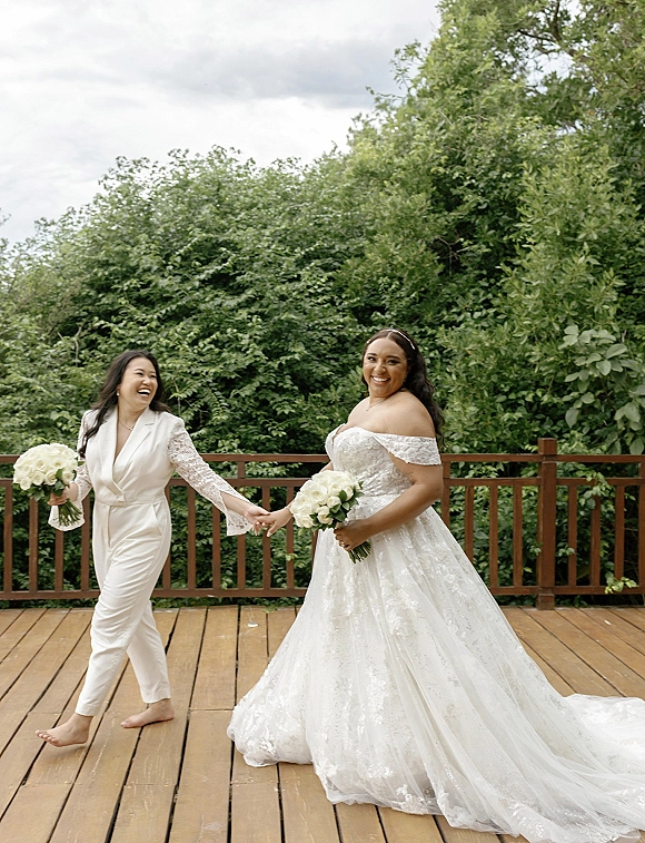 Bride portrait of two brides holding hands, one in off-shoulder lace dress and one in white jumpsuit on a wood deck with trees under cloudy sky